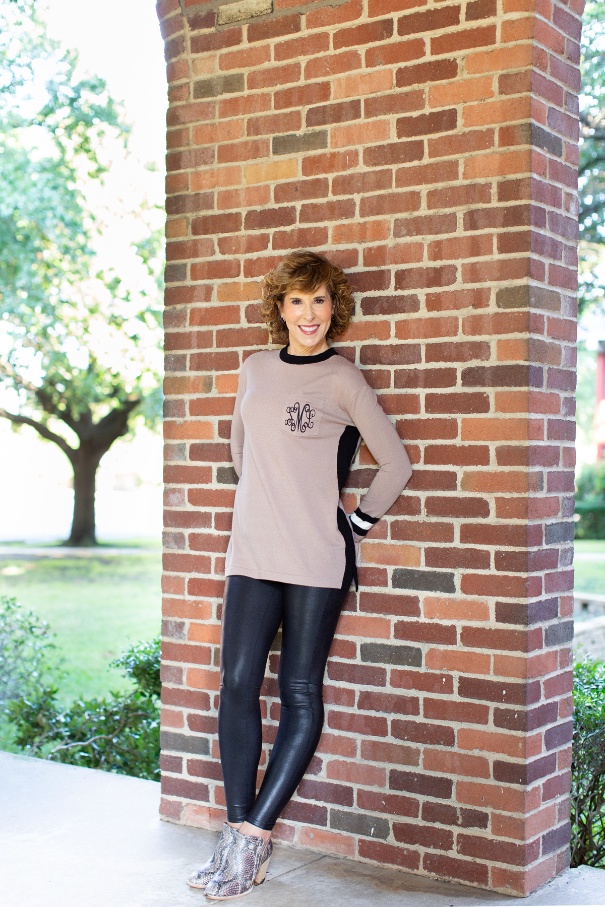 woman in tan monogrammed sweater standing in front of a brick wall