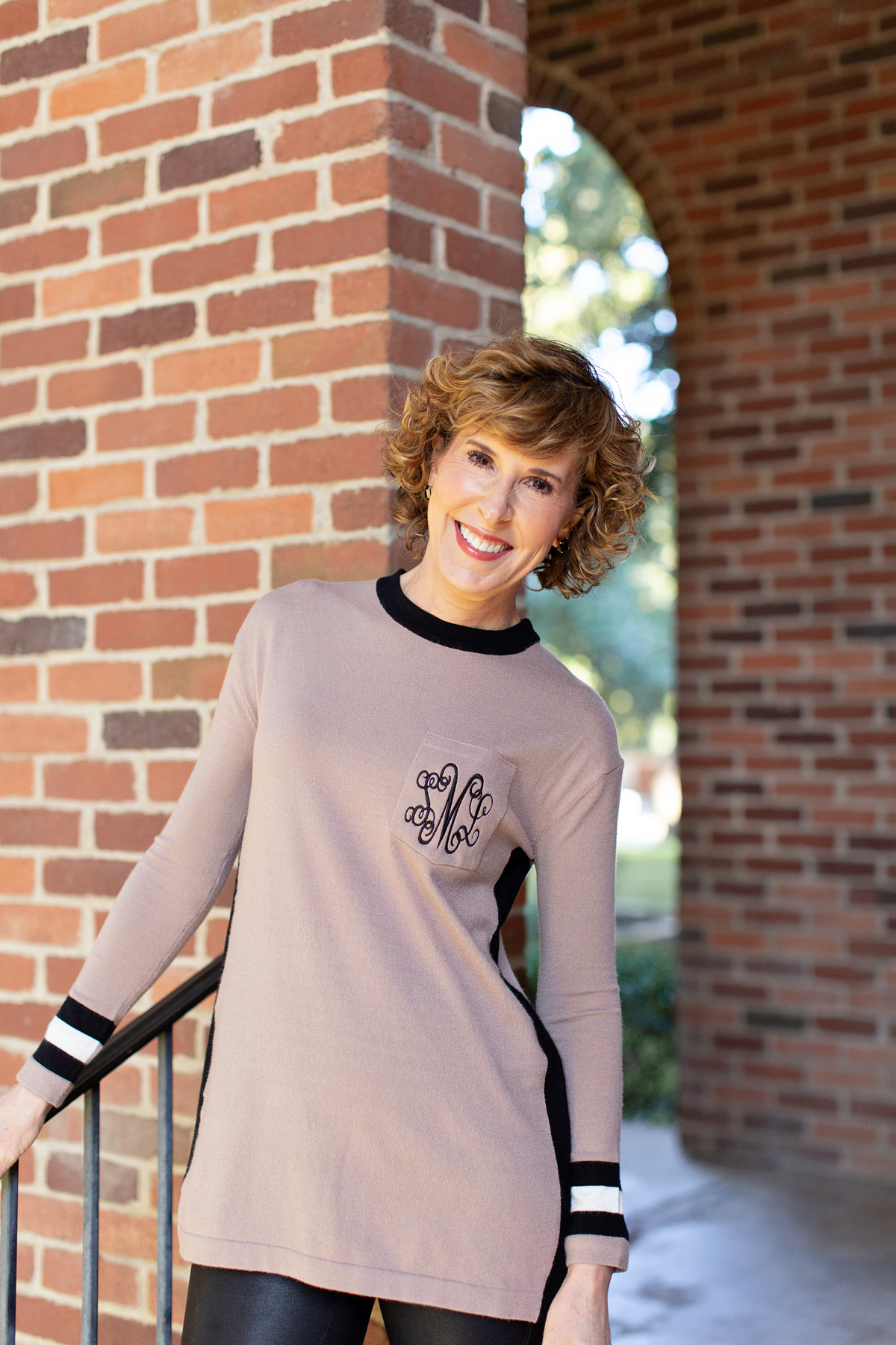 woman in tan monogrammed sweater standing in front of a brick wall