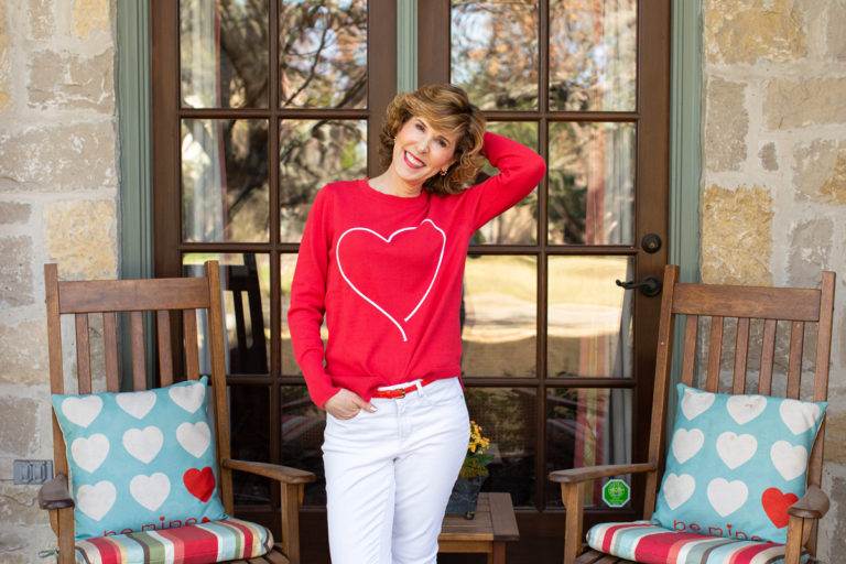 woman in red heart sweater, white jeans, and red sneakers on a porch