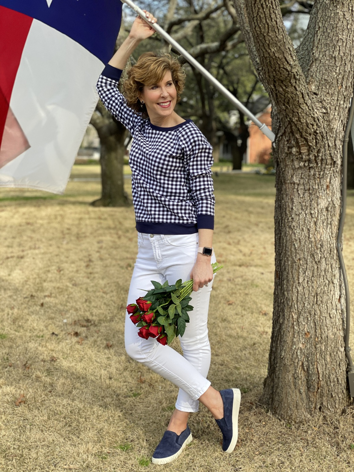 woman over 50 wearing navy and white gingham sweatshirt and white jeans standing by a texas flag and holding red roses