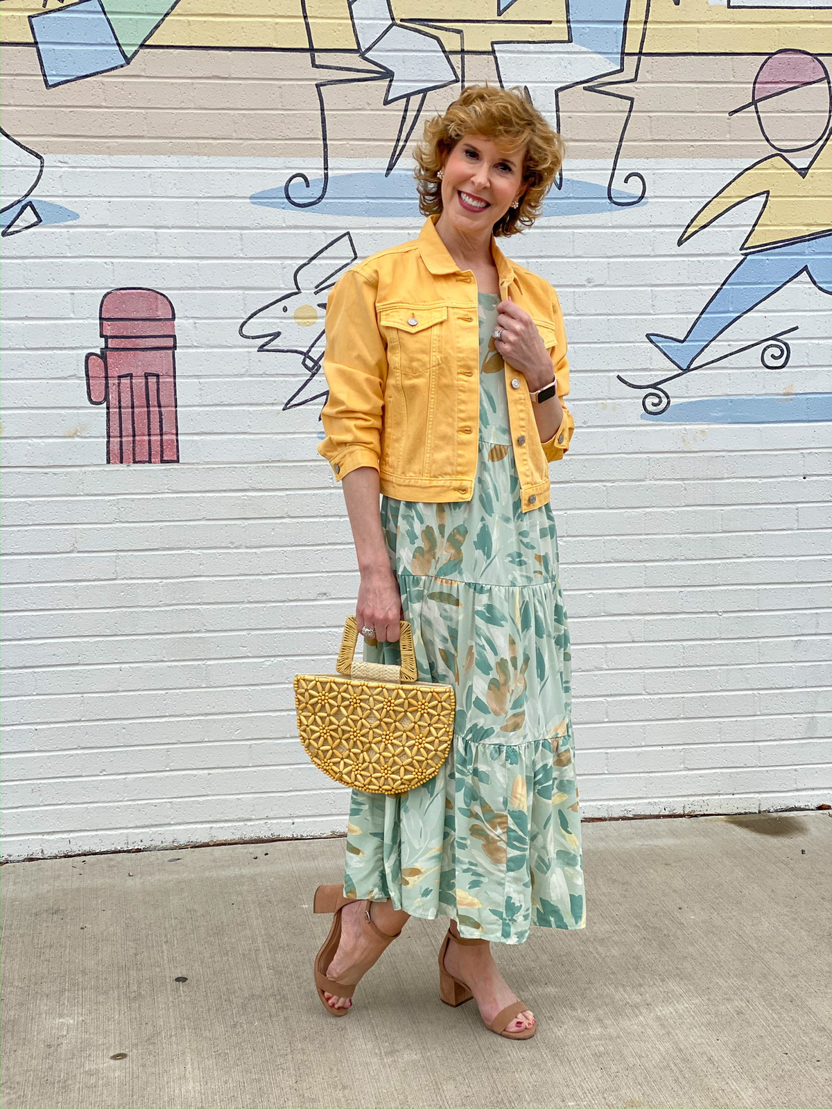 woman in green print dress and yellow jacket in front of a mural