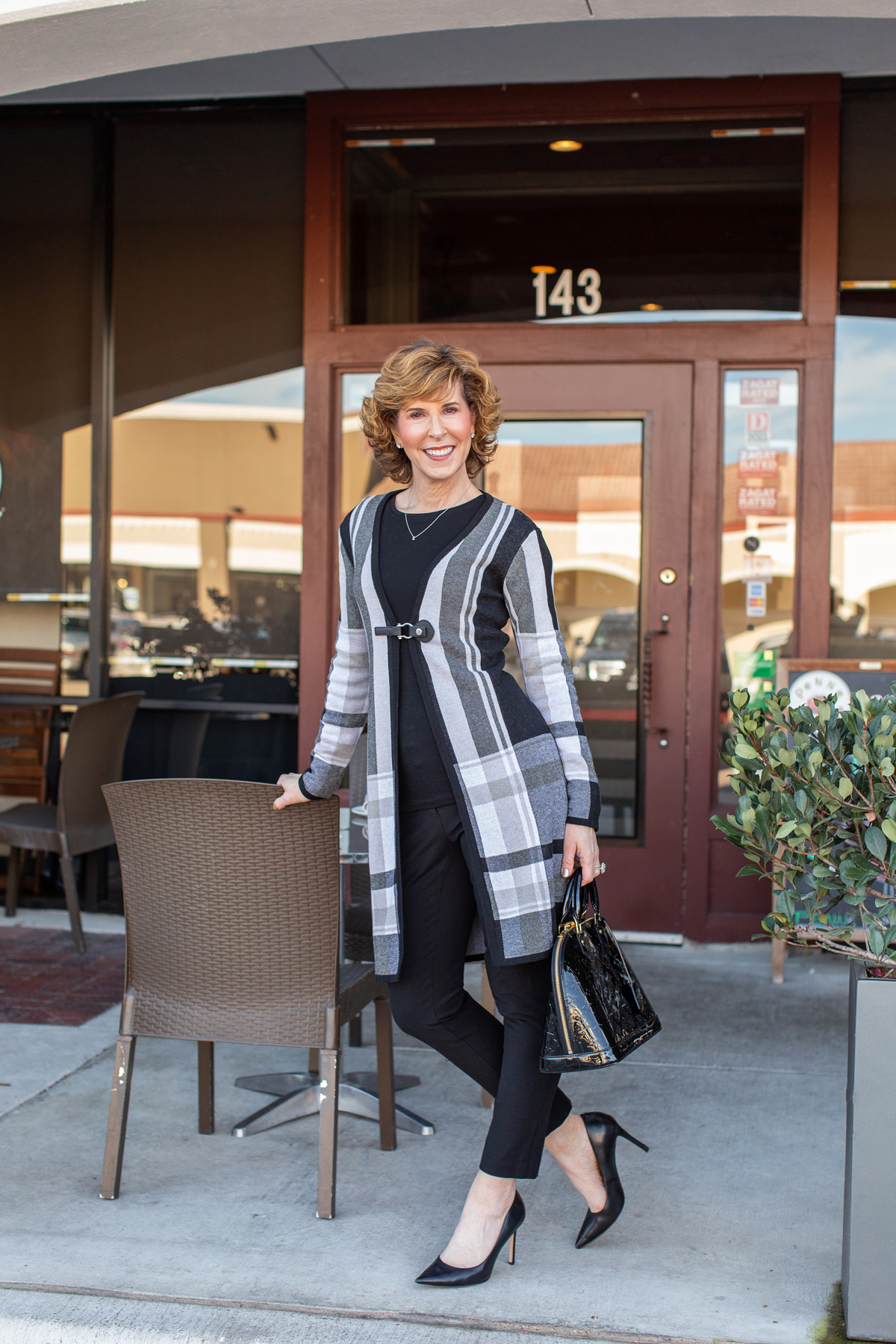 woman over fifty wearing a black plaid coat and black pants standing in front of a restaurant