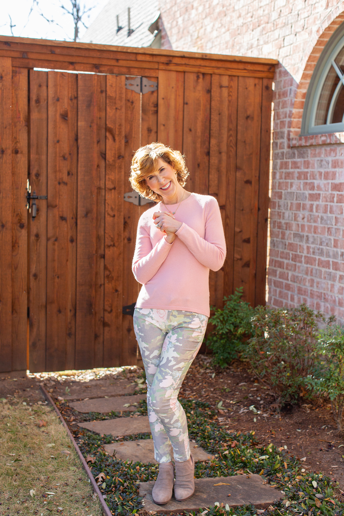 woman wearing pink sweater and floral camo jeans in front of a brown fence