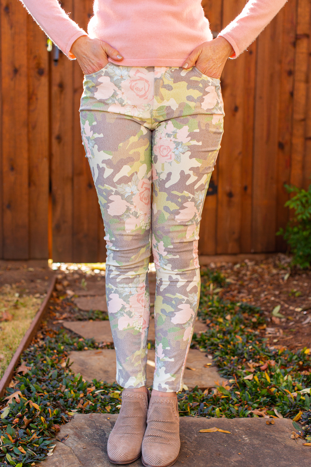 woman wearing pink sweater and floral camo jeans in front of a brown fence