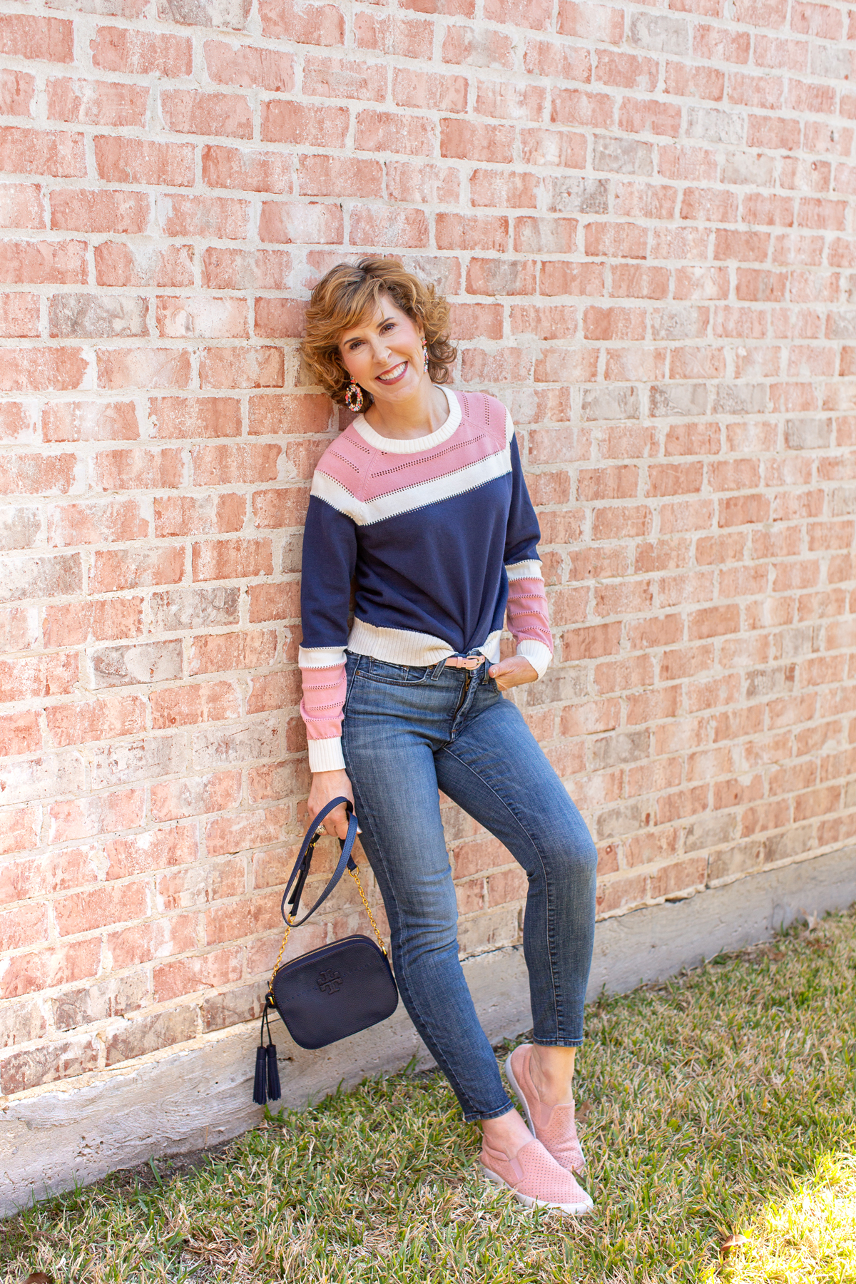 woman wearing blue and pink sweater and jeans leaning against a brick wall