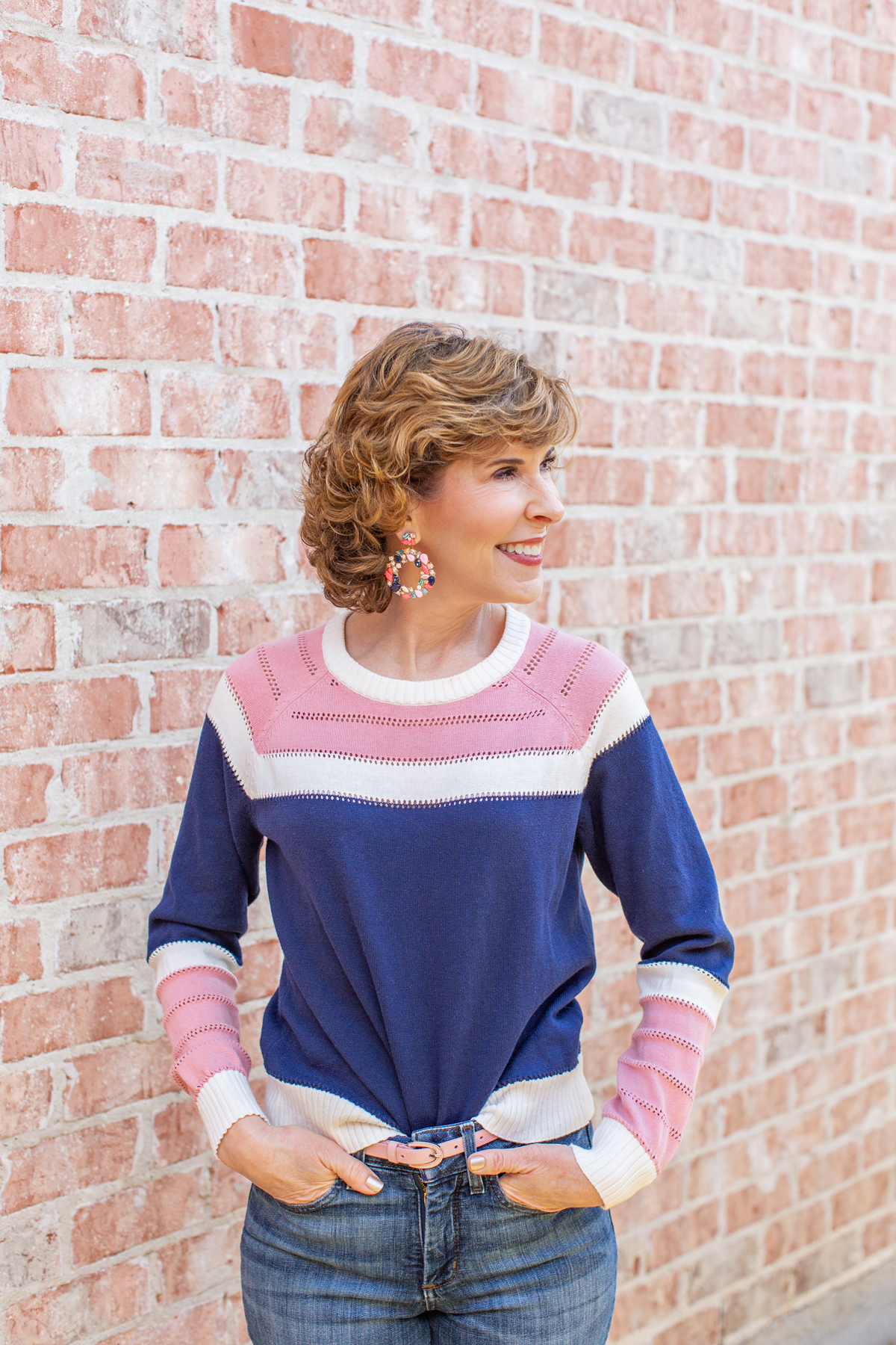 woman wearing blue and pink sweater and jeans leaning against a brick wall
