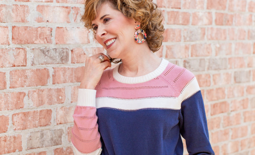 woman wearing blue and pink sweater and jeans leaning against a brick wall