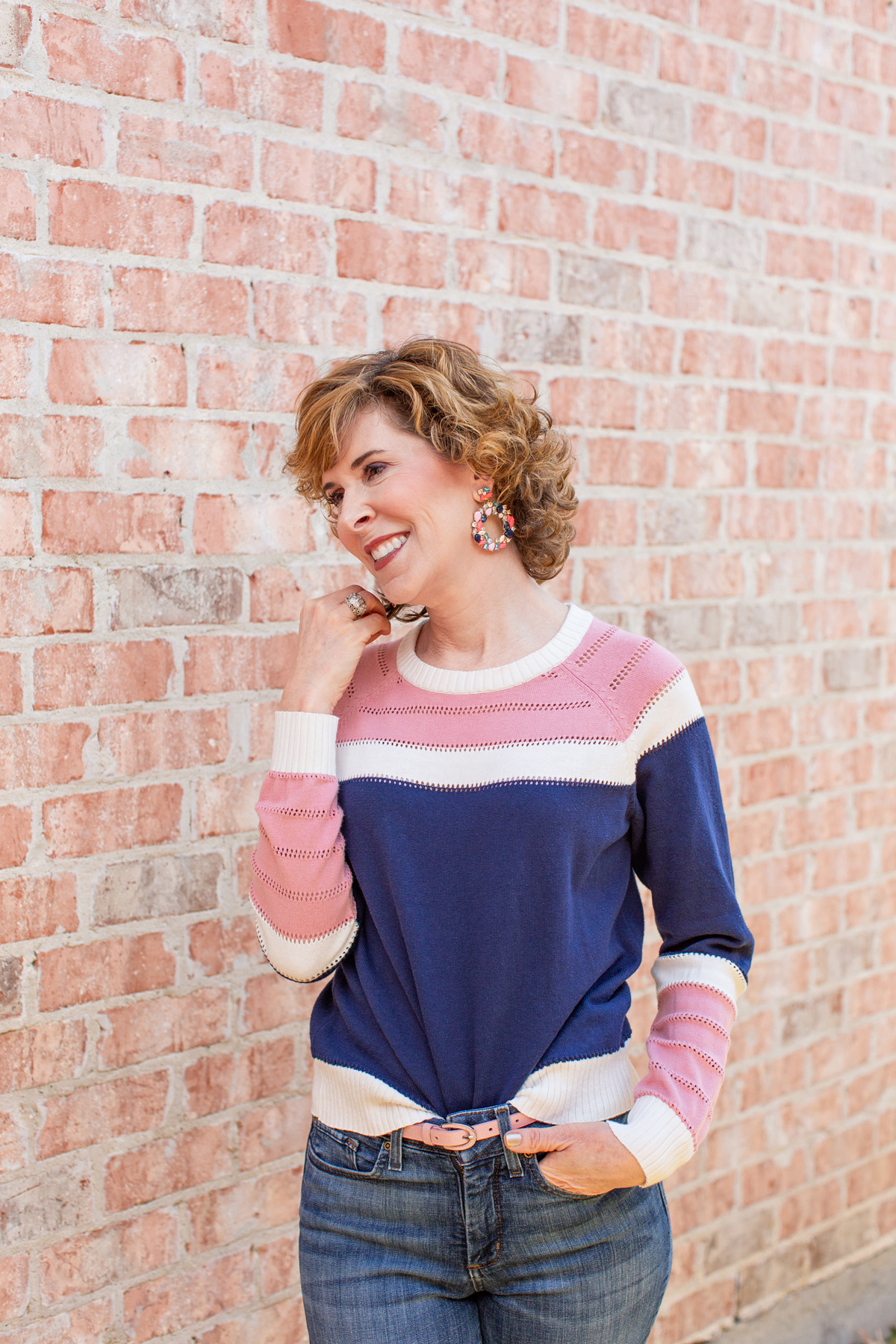 woman wearing blue and pink sweater and jeans leaning against a brick wall