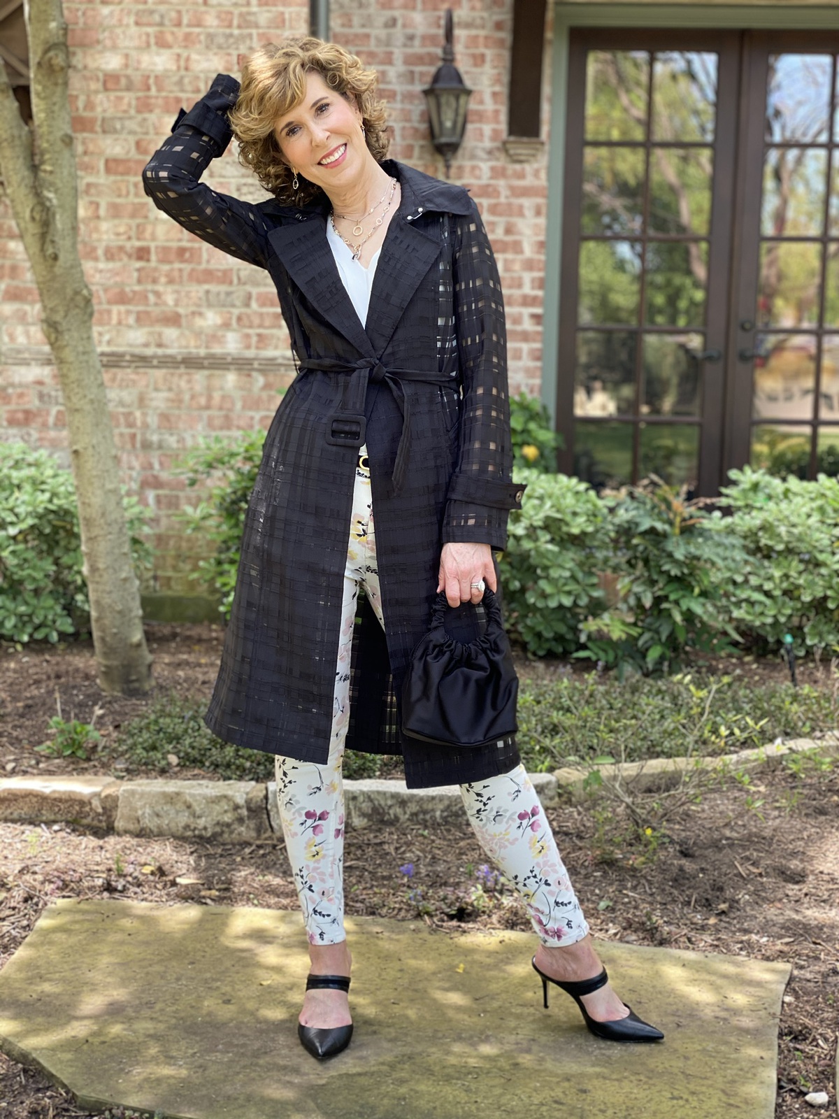 woman in black trench coat and floral pants standing in front of house