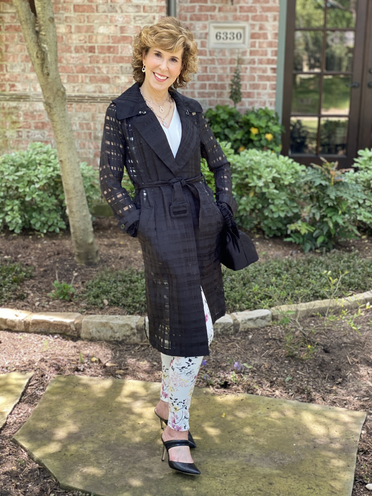 woman in black trench coat and floral pants standing in front yard