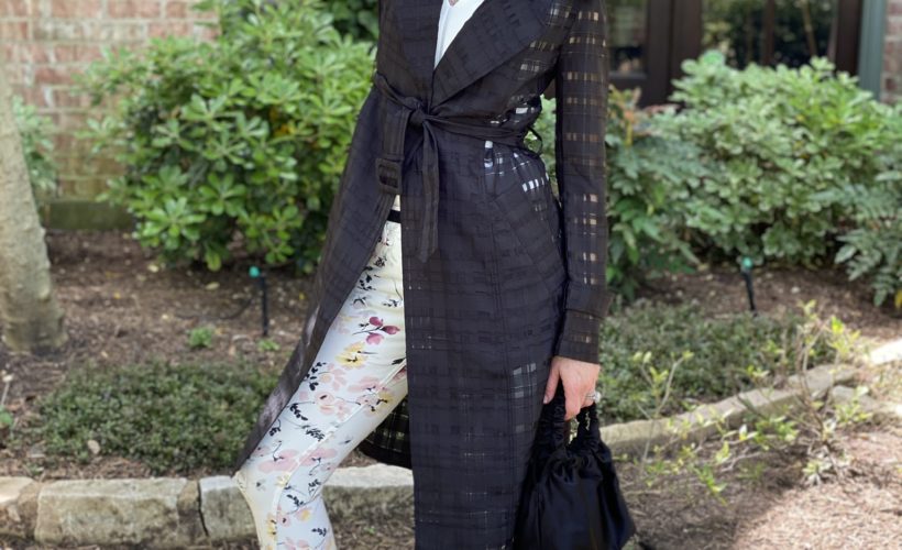 woman in black trench coat and floral pants standing in front of house