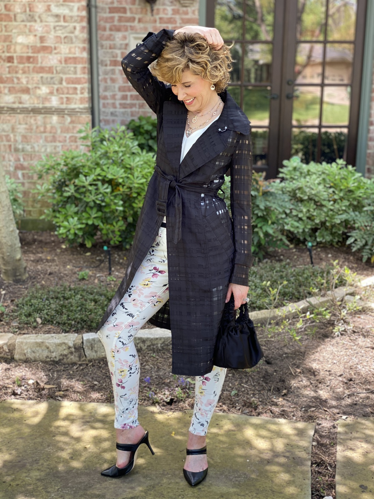 woman in black trench coat and floral pants standing in front of house