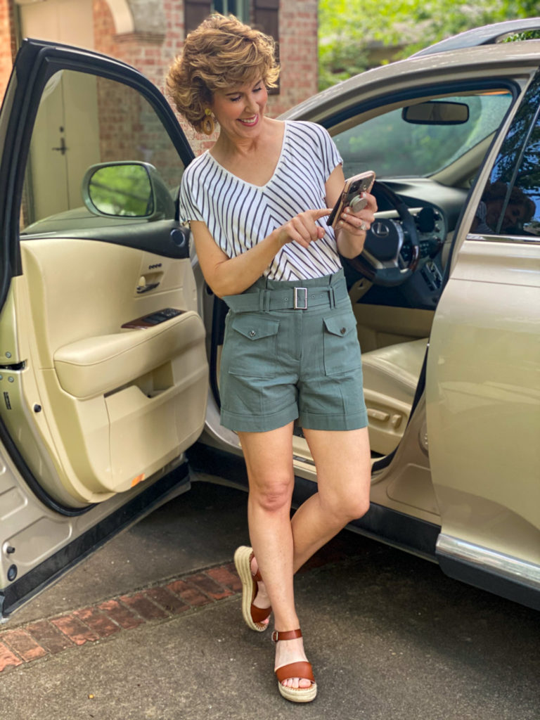 woman in green striped shirt and green shorts standing by car looking at cell phone