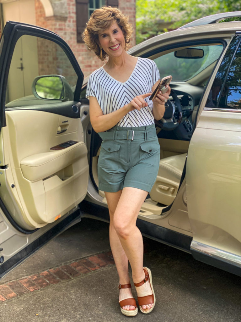 woman in green striped shirt and green shorts standing by car looking at cell phone