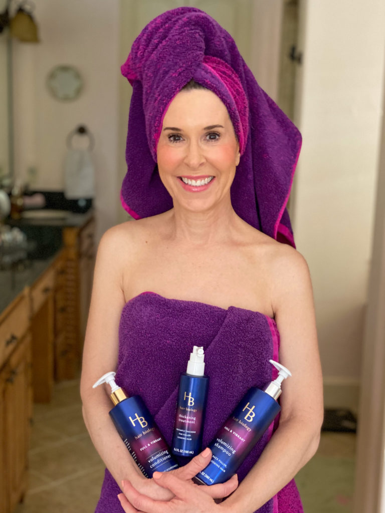 woman with hair over 50 dressed in a purple towel and holding hair biology volumizing treatement and a pink blow dryer