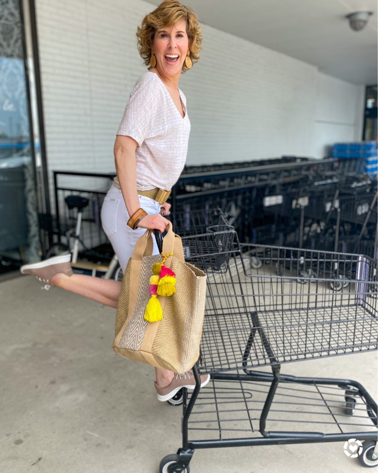 woman wearing tan tee and white shorts playfully standing on back of grocery cart doing weekend shopping