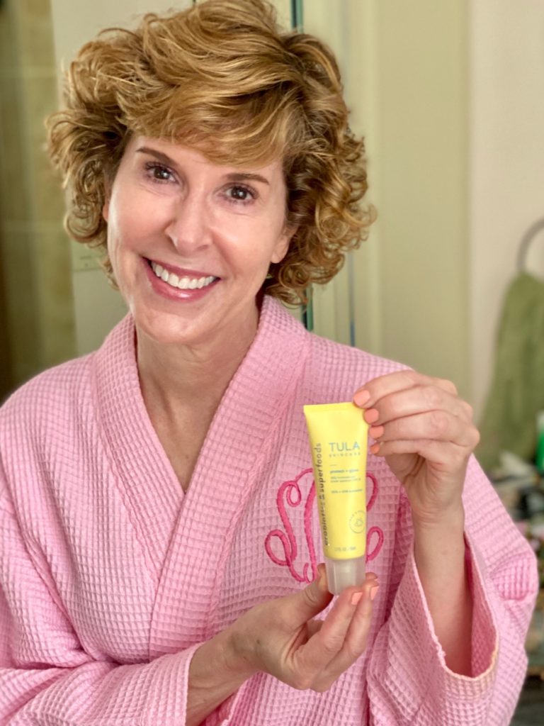 woman dressed in pink robe displaying tula protect and glow sunscreen