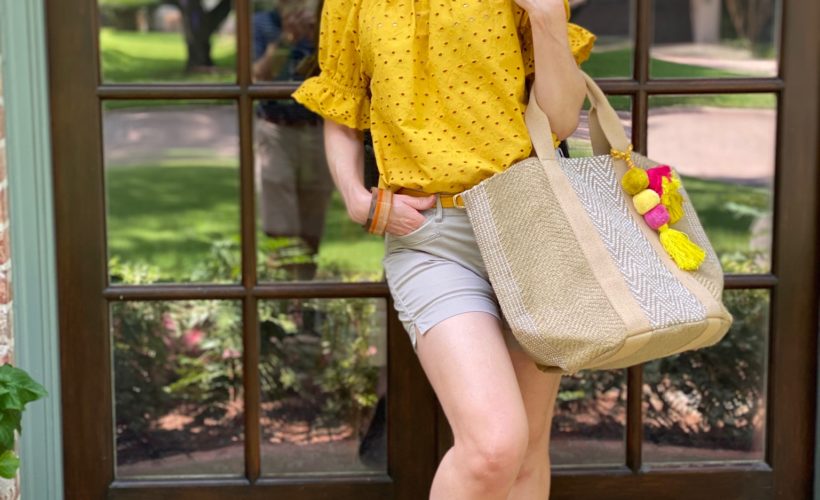 woman dressed in yellow eyelet top and tan shorts carrying burlap tote with tassels standing on porch in front of french doors