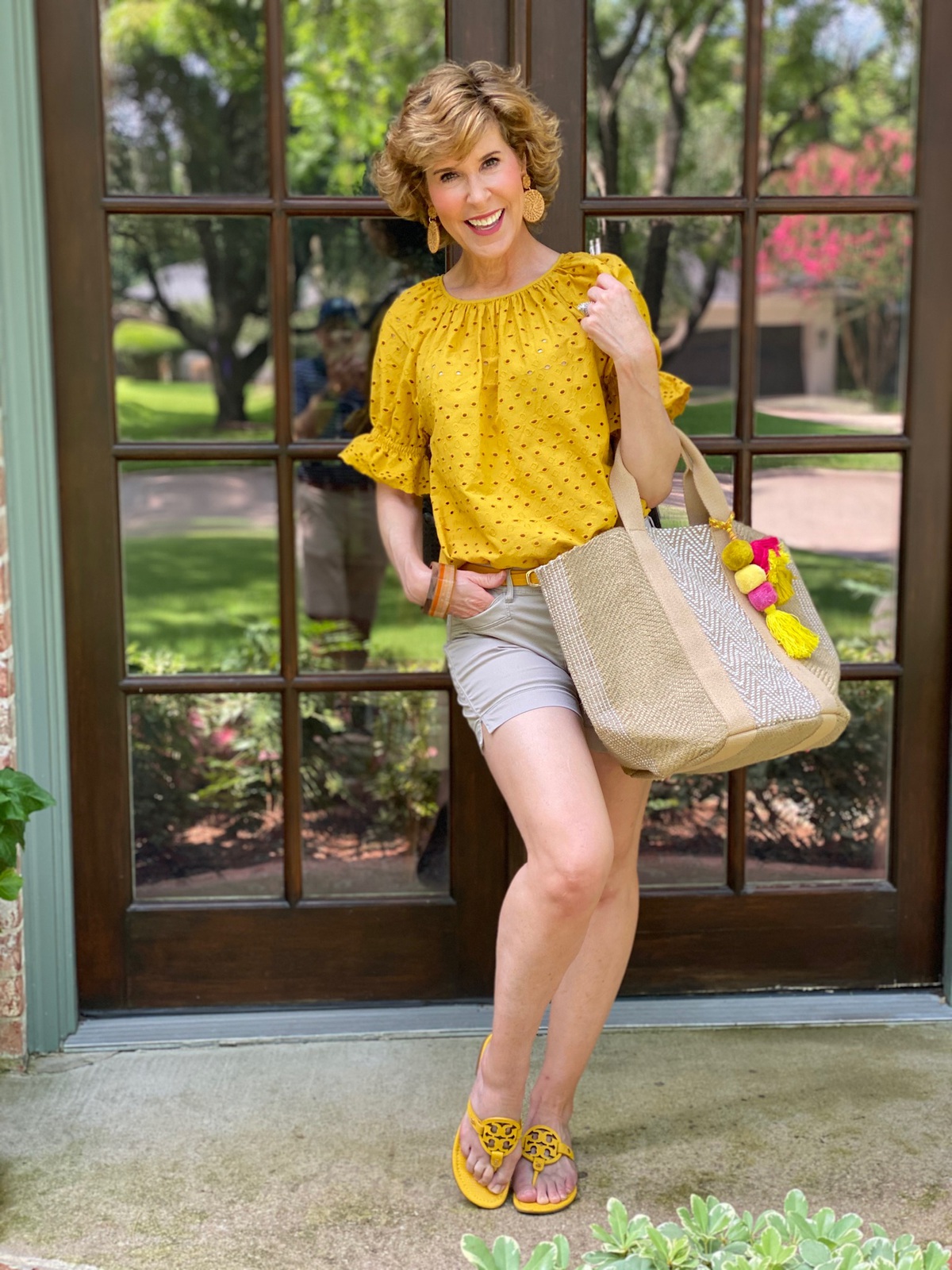 woman dressed in yellow eyelet top and tan shorts carrying burlap tote with tassels standing on porch in front of french doors