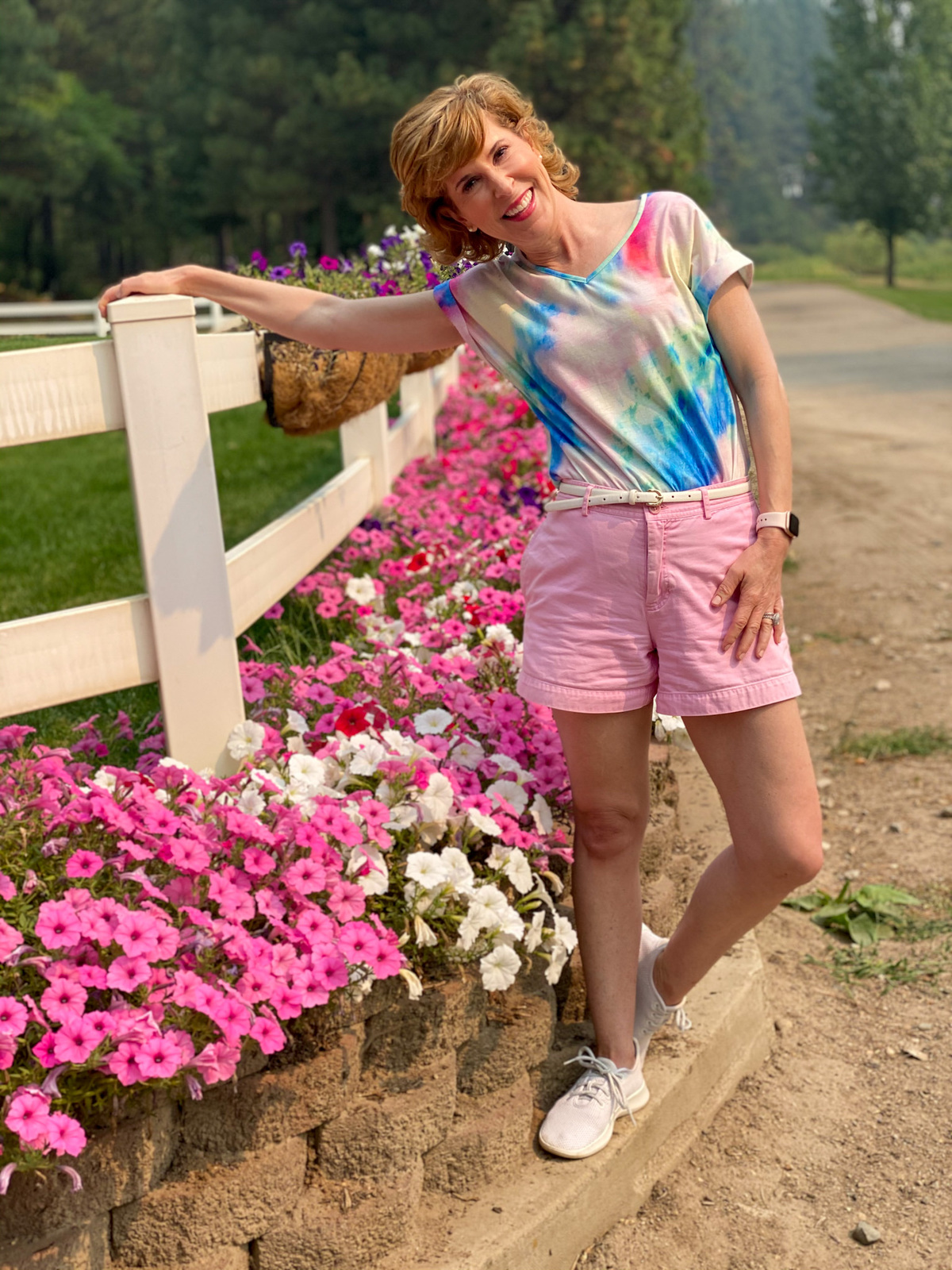 woman wearing tie dye shirt and pink shorts standing by white fence and flowers