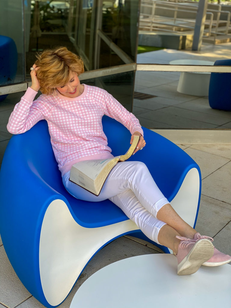 woman in pink gingham sweatshirt, white jeans, pink sneakers holding her Bible and sitting in a blue chair