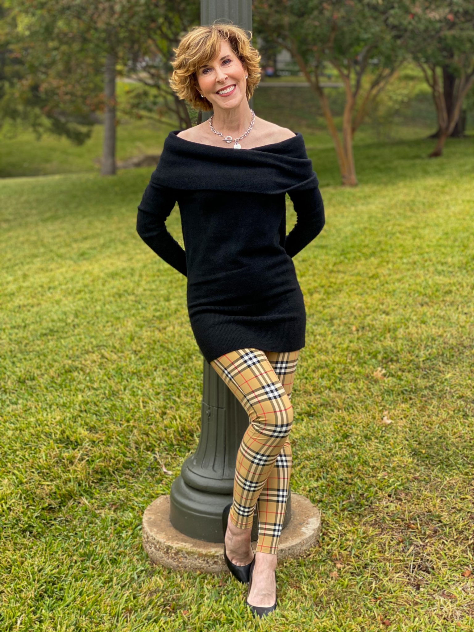 woman over 50 wearing a black off the shoulder sweater and burberry leggings and black pumps leaning back against a green lamppost