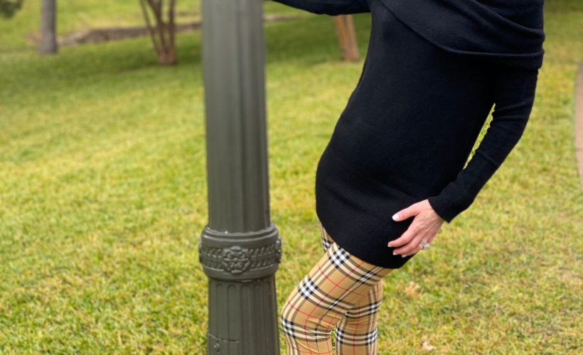 woman over 50 wearing a black off the shoulder sweater and burberry leggings and black pumps with one hand on a lamppost leaning away from it