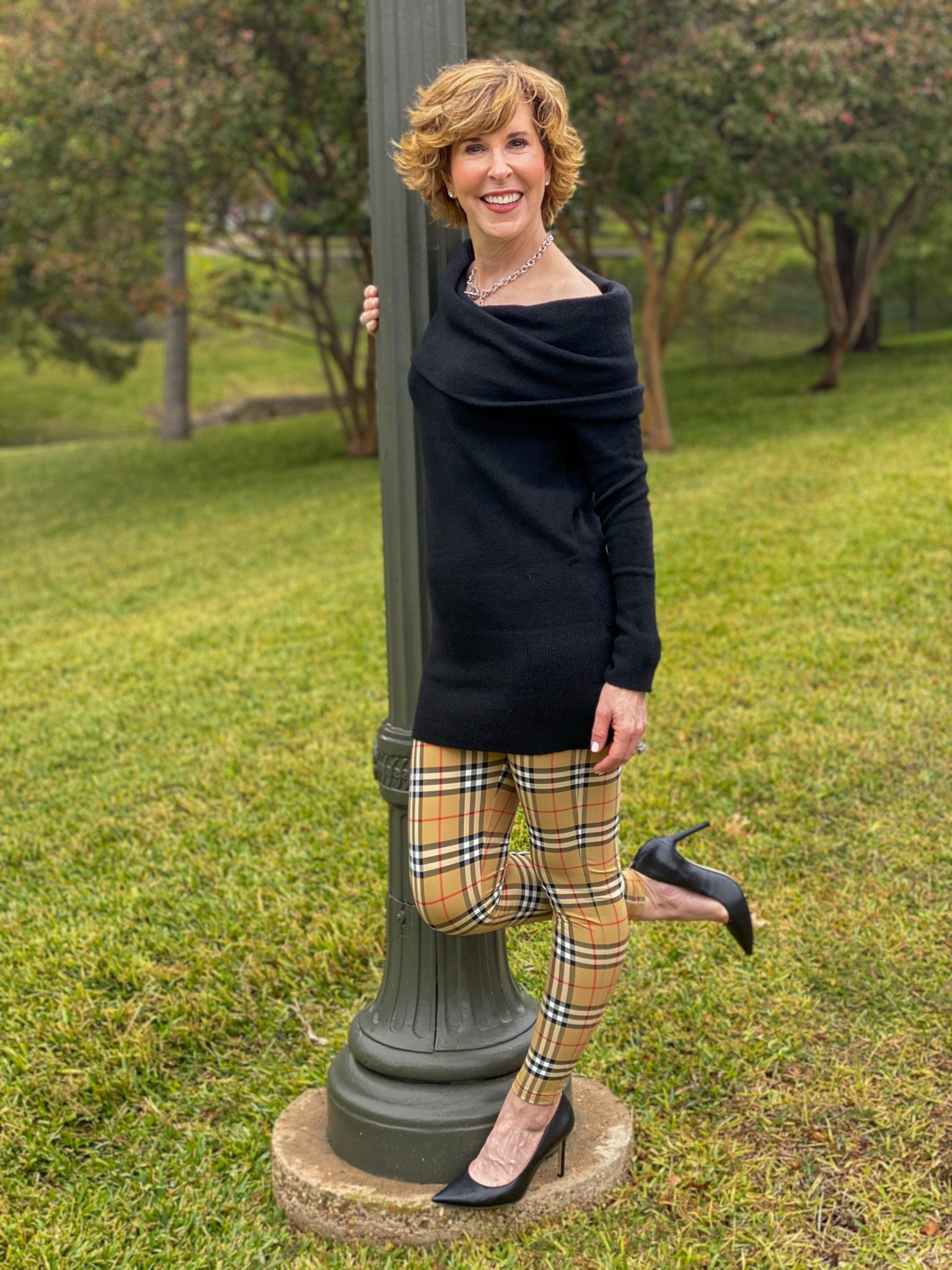 woman over 50 wearing a black off the shoulder sweater and burberry leggings and black pumps standing next to a green lamppost with one leg up in the air