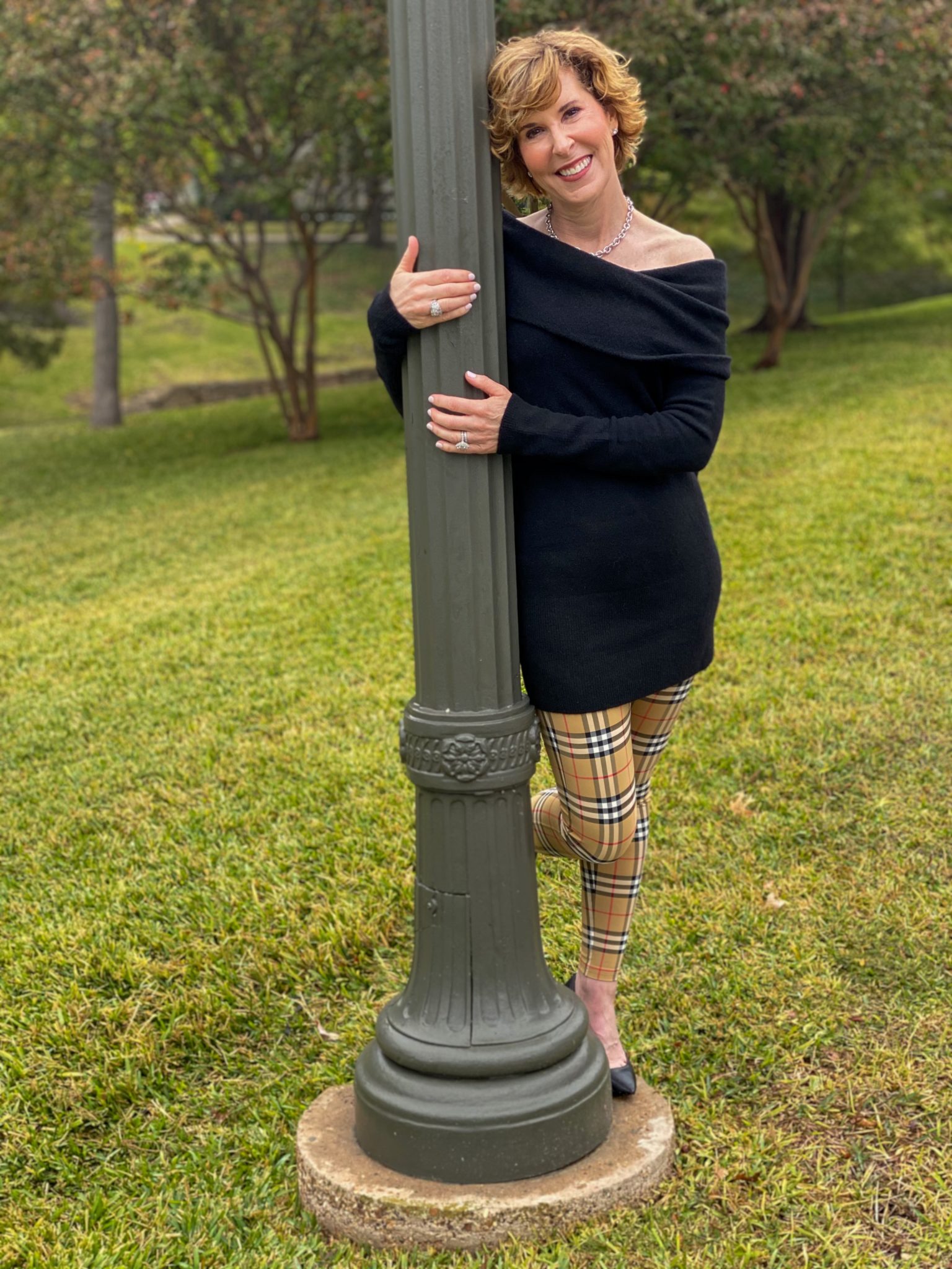 woman over 50 wearing a black off the shoulder sweater and burberry leggings and black pumps with her arms around a lamppost