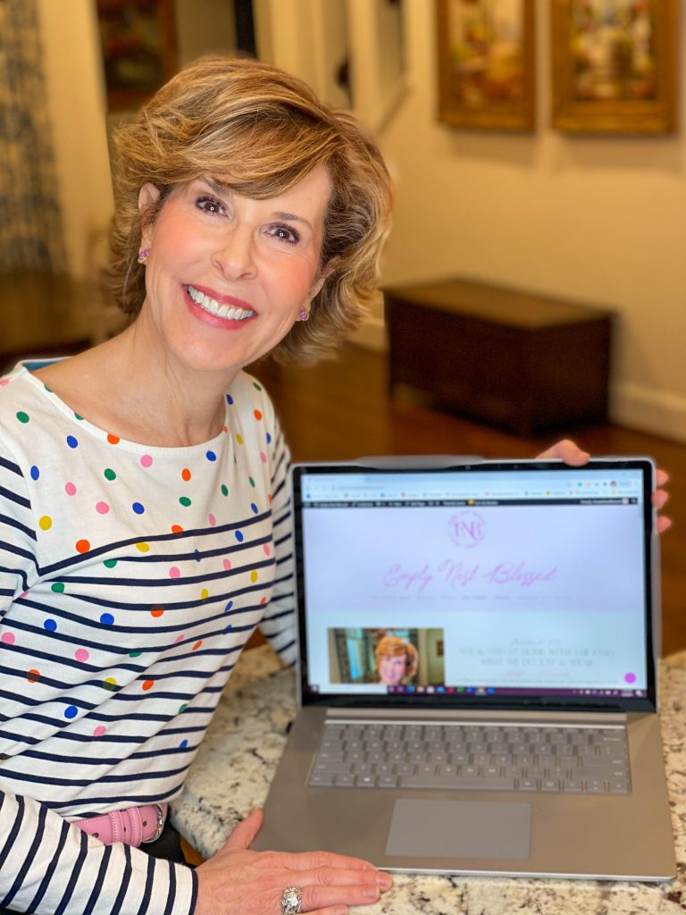 woman over 50 dressed in boden multi colored polka dot and striped Breton shirt sitting next to a computer displaying new empty nest blessed website