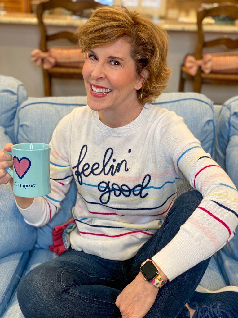 woman wearing walmart feelin' good striped time and tru sweater sitting on a blue sofa holdng a coffee cup talking about skin care over 50