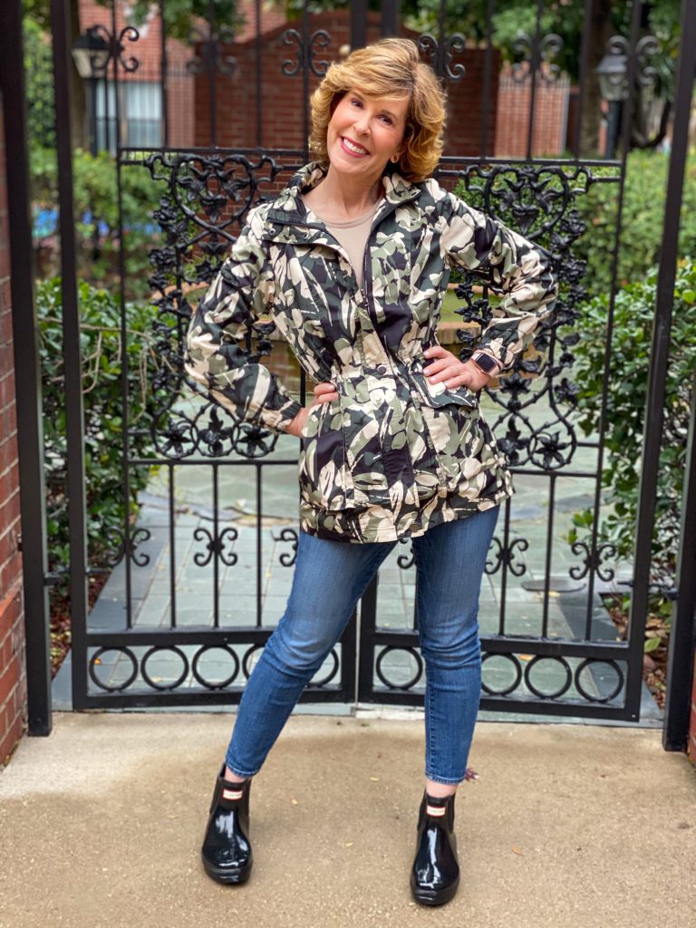woman over 50 posing in front of a black iron gate wearing chico's floral camo raincoat and black hunter chelsea boots with jeans