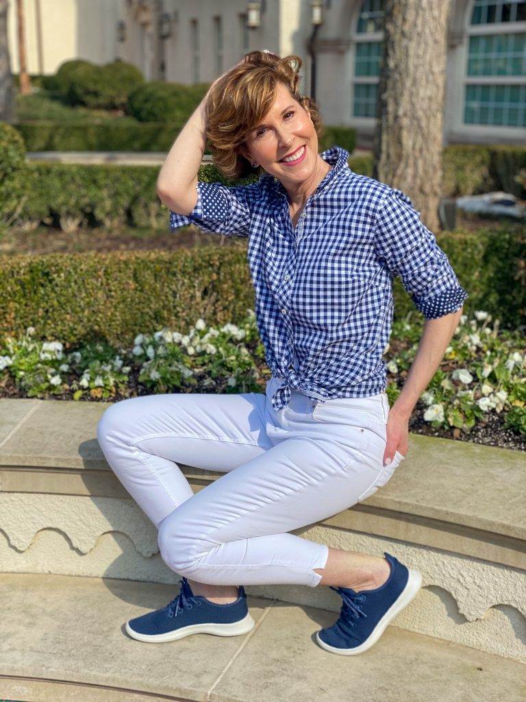 woman wearing talbots blue and white gingham shirt and white jeans with navy allbirds tree runner sneakers sitting on a wall