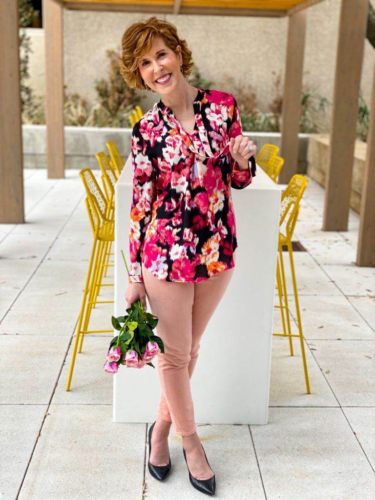 Woman over 50 wearing chico's floral tie-neck blouse with azalea jeggings and black pumps holding pink roses standing in front of a long table with yellow chairs