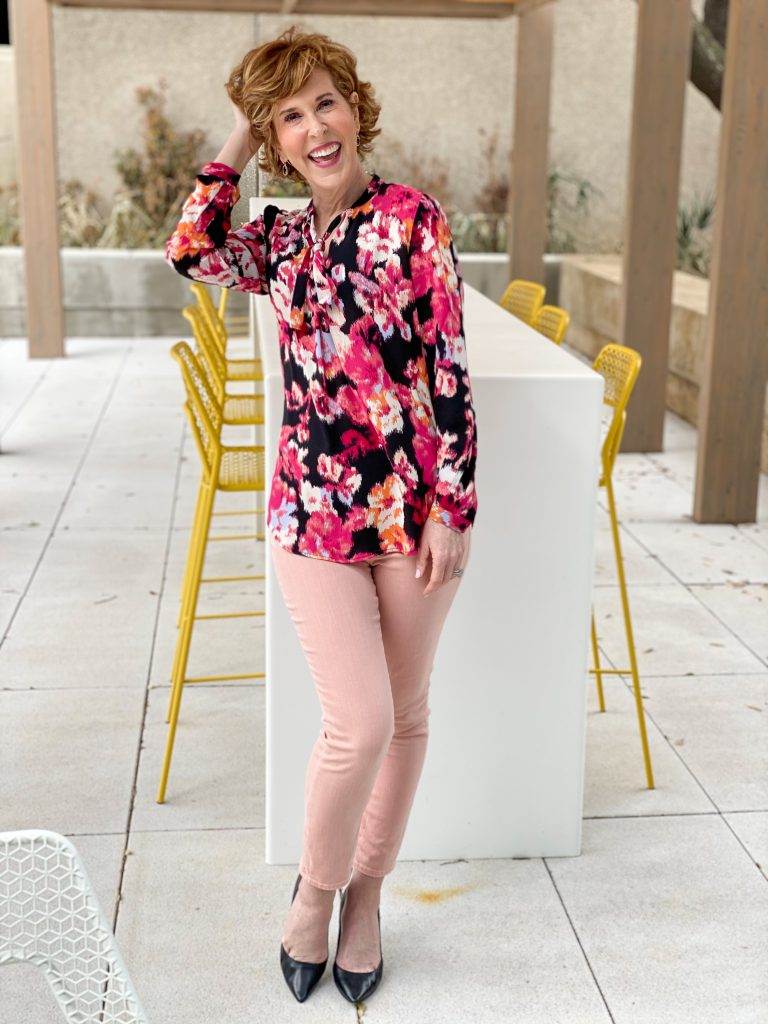 Woman over 50 wearing chico's floral tie-neck blouse with azalea jeggings and black pumps holding pink roses standing in front of a long table with yellow chairs