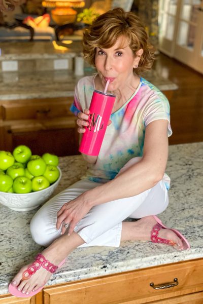 woman wearing tie dye shirt and white jeans with pink sandals holding a Marley Lilly pink tumbler sitting on a counter