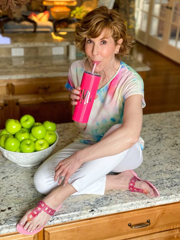woman wearing tie dye shirt and white jeans with pink sandals holding a Marley Lilly pink tumbler sitting on a counter