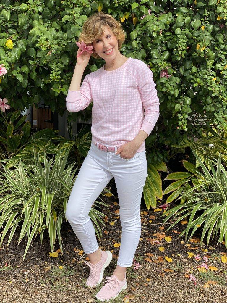 woman wearing draper james pink and white gingham sweatshirt and white jeans standing in front of bushes with a flower in her hair