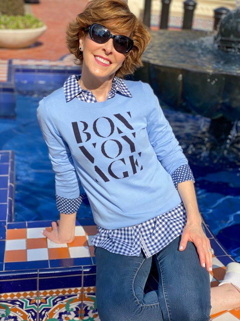 over 50 woman in sunglasses wearing Talbots blue bon voyage sweater over a blue and white gingham button down with jeans and sneakers sitting on the edge of a colorful fountain