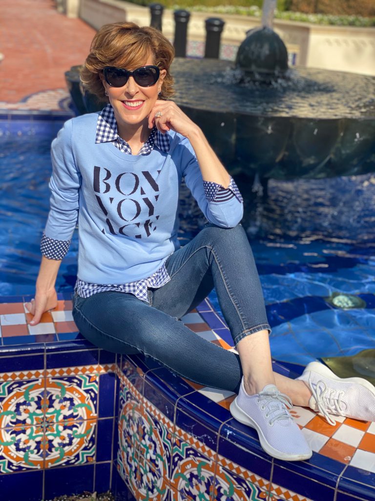 over 50 woman in sunglasses wearing Talbots blue bon voyage sweater over a blue and white gingham button down with jeans and sneakers sitting on the edge of a colorful fountain