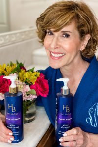 woman in blue robe with hair biology products and a bouquet of flowers behind her