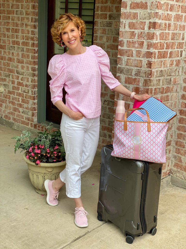 woman wearing pink eyelet top and white jeans standing by a suitcase getting ready for travel
