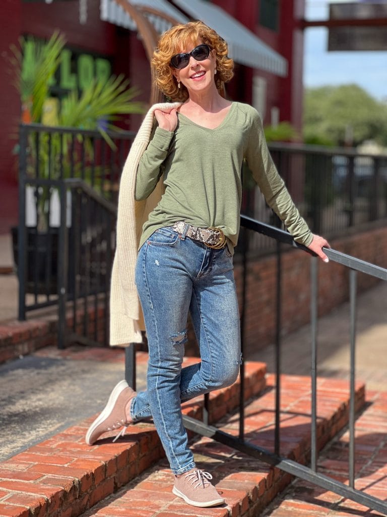 woman over 50 wearing cabi recline tee, cinch skinny jeans and holding the book club cardigan standing on a brick staircase by a black railing