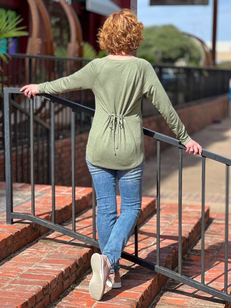 woman over 50 wearing cabi recline tee and cinch skinny jeans standing on a brick staircase outside with her back to the camera