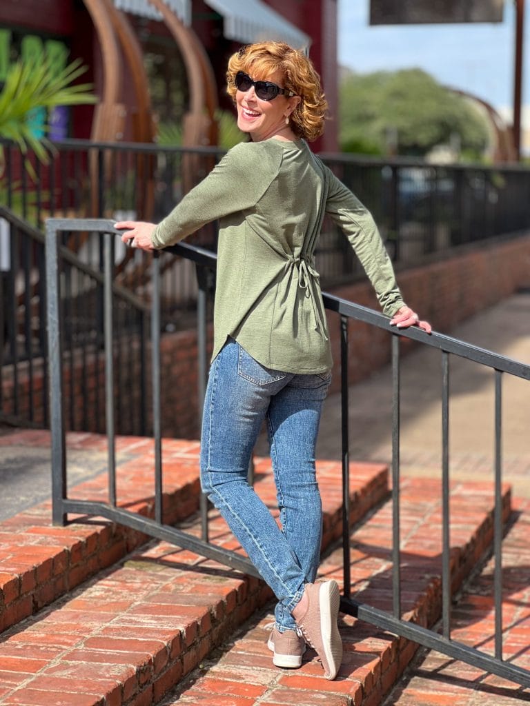 woman over 50 looking backwards over her shoulder and wearing cabi recline tee and cinch skinny jeans sittring on a brick staircase outside