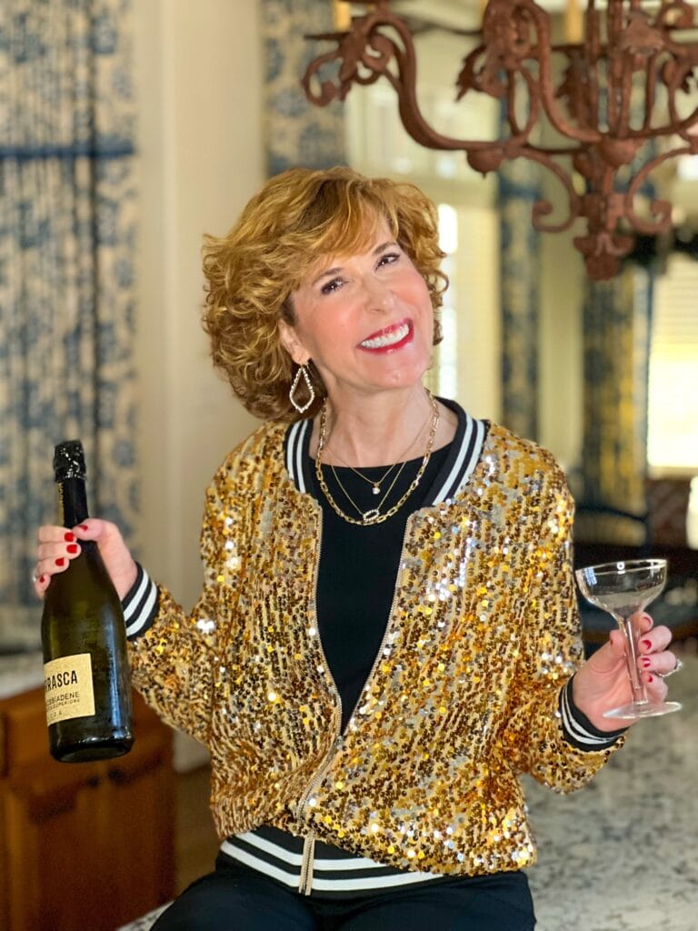 woman wearing sequin jacket holding a champagne bottle and glass celebrating nye at home