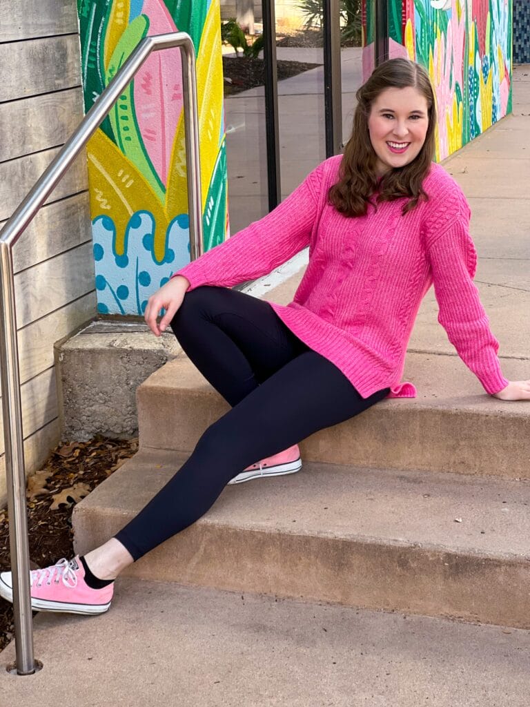 20 something brunette girl wearing cabi elsa pullover and black leggings with pink converse sneakers sitting on a staircase