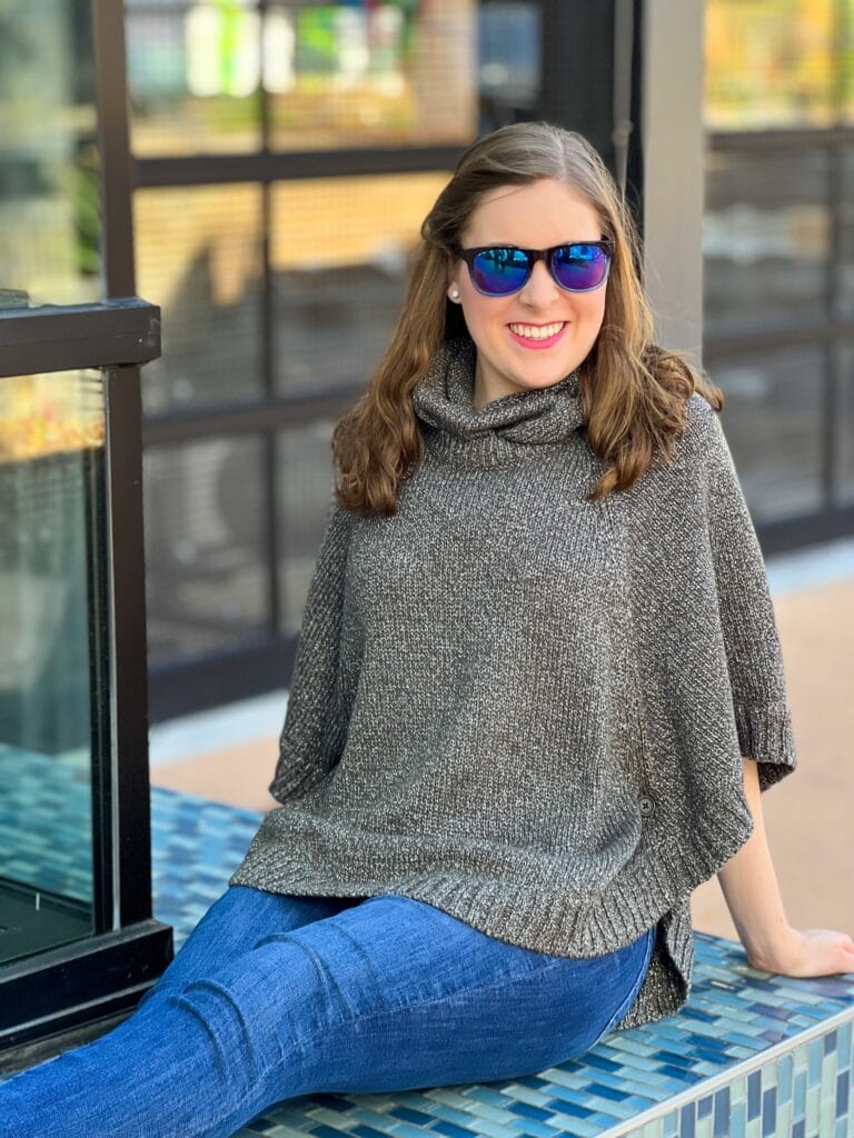 20 something girl wearing cabi shimmer poncho jeans and sunglasses sitting on blue tiled bench