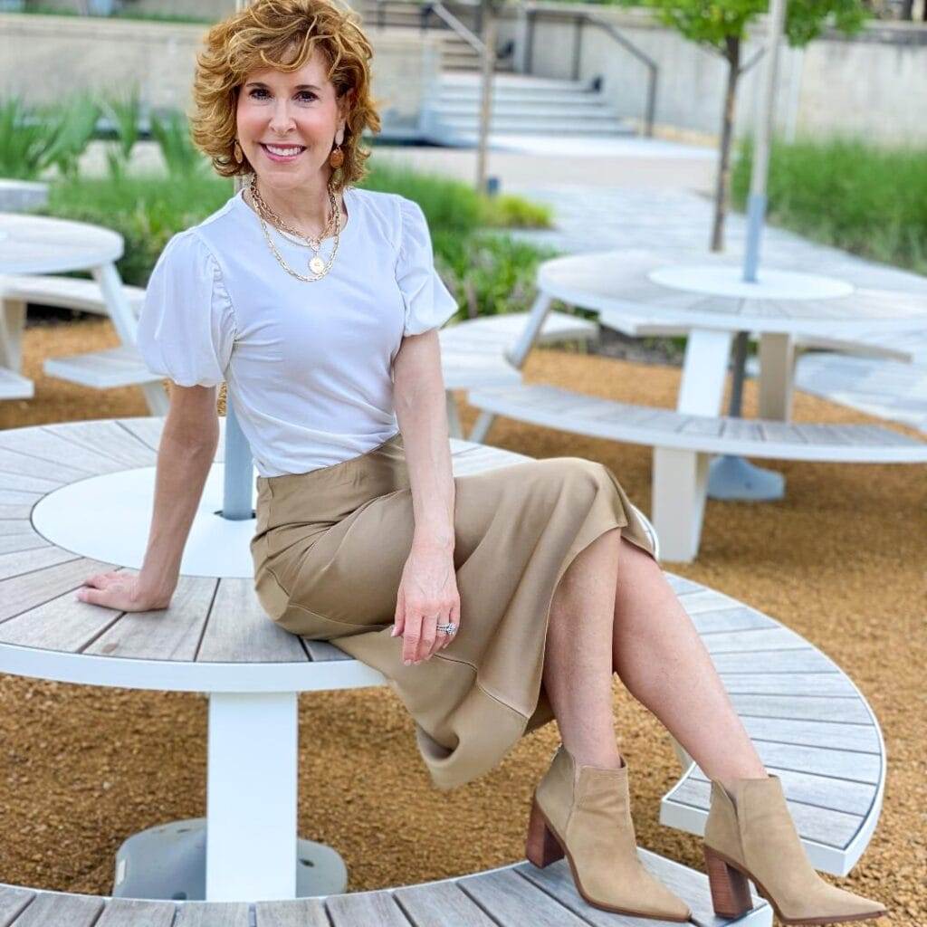 woman over 50 wearing a white puff sleeve tee and jcrew taupe slip skirt with taupe booties sitting on top of a round white picnic table