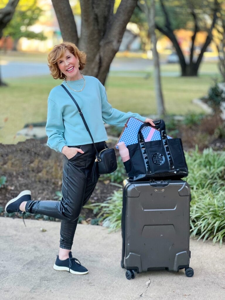 woman over 50 wearing light blue sweatshirt and spanx faux leather jogger with allbirds black wool runners posing on a driveway by a suitcase and tory burch tote bag